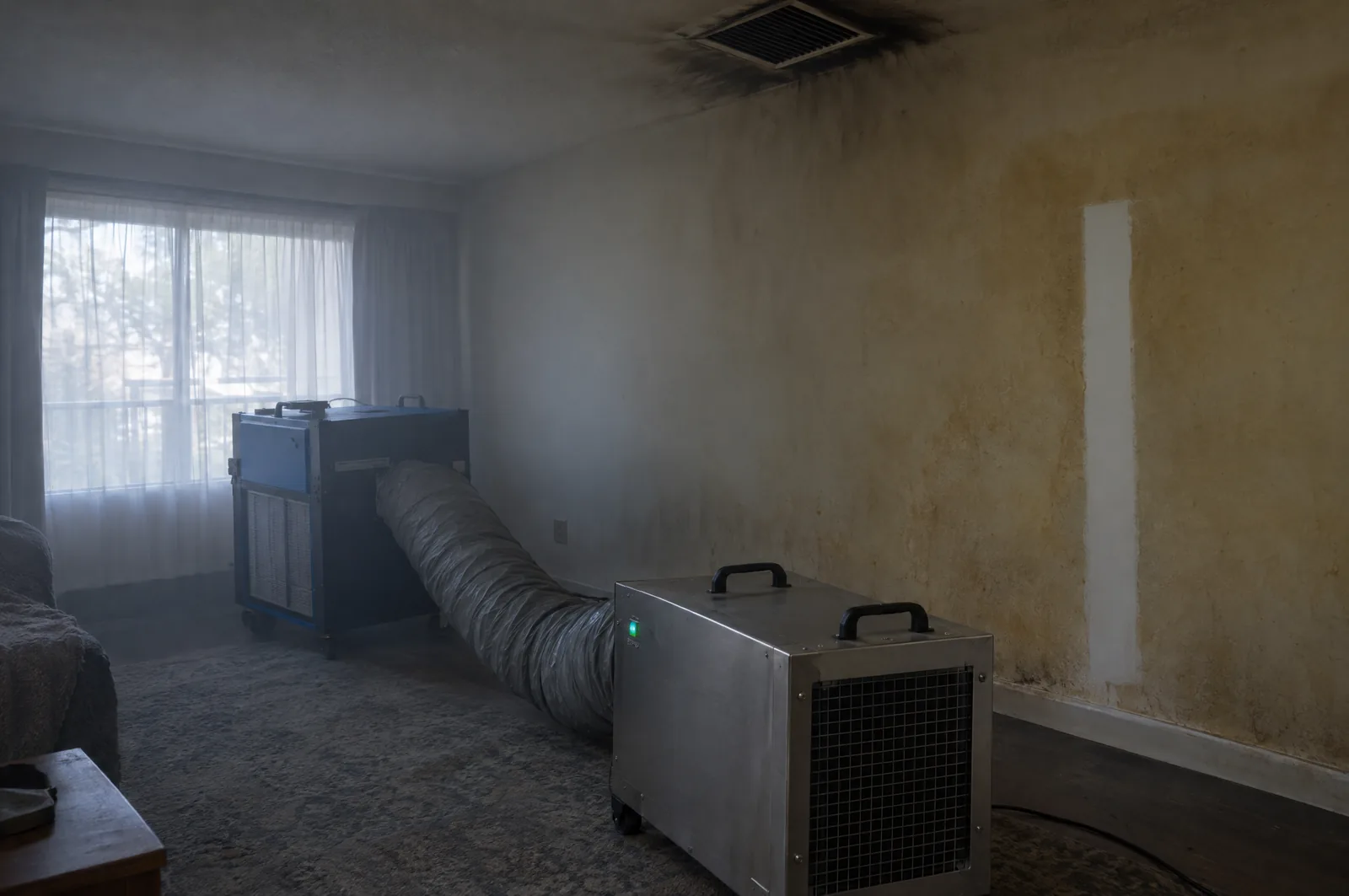 Hydroxyl generator and HEPA air scrubber with ducting running in a smoke-damaged room, with soot staining around the ceiling vent and a clean wipe test strip on the wall